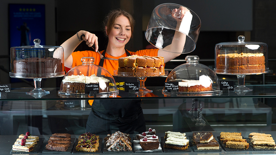 A server at our café uses a cake slice to serve a segment of Victoria Sponge. On the counter alongside the Victoria Sponge are other cakes, both on top and inside the glass display.