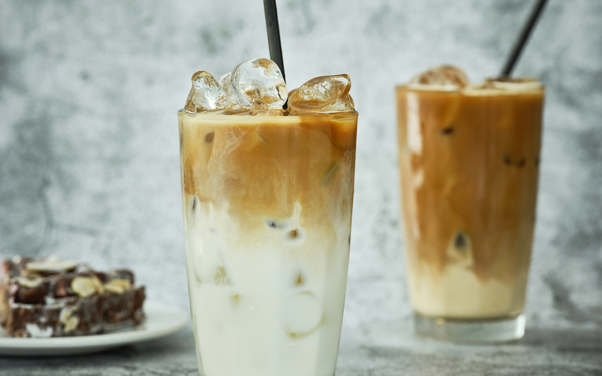 Two iced coffees with black paper straws alongside a Rocky Road brownie, which is on a small white plate. Both items are on a grey stone textured floor with the same pattern background.