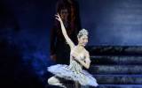 A ballerina in a sparkling silver tutu and tiara balances en pointe with one leg raised, elegantly posing against a dark stage with figures in the background.