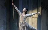 A ballerina dressed as Cinderella gracefully leaps with one leg extended behind her, holding a broomstick aloft in a dimly lit setting with an old wooden wardrobe in the background.