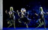 Three dancers in elaborate frog costumes, wearing formal coats and scarves, leap mid-air against a dark, starry backdrop.